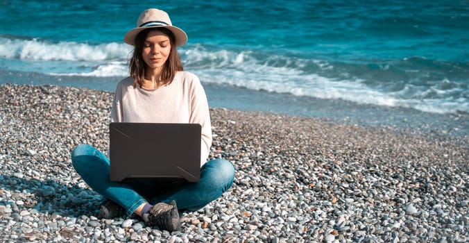 A young girl in a light hat and casual sweater lies on the beach by the sea with a laptop on a sunny day, works, studies, buys tickets during trip, a woman rests on vacation and types on the keyboard.