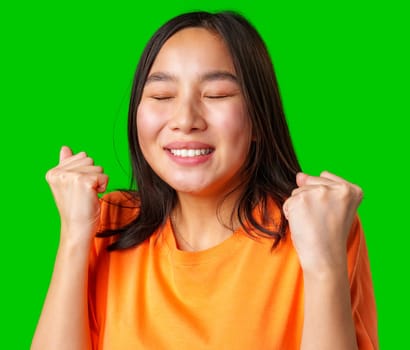 A young woman in an orange shirt expresses joy and triumph with her eyes closed and fists raised. The bright green backdrop enhances her delightful enthusiasm.