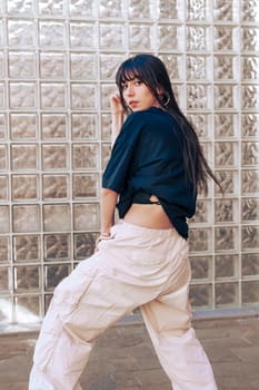 A young woman showcases her fashion sense in an urban environment, wearing a trendy outfit with loose-fitting pants. The glass wall adds a modern touch to the scene.