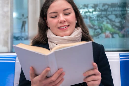 Young woman in a black coat and beige scarf sitting inside a modern subway train, reading a book with focus. She enjoys a quiet moment during her commute in the city.