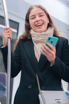 Cheerful young woman in a black coat and beige scarf standing inside a modern subway train, holding a smartphone and gripping a metal pole for support. She smiles brightly during her commute.