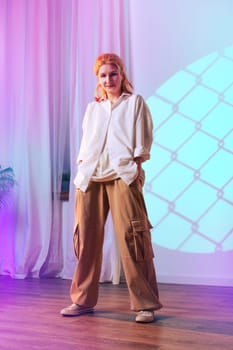 A woman stands confidently, wearing a loose white shirt and tan cargo pants. She poses in an indoor space highlighted with pastel lighting and decorative curtains, creating a trendy atmosphere.