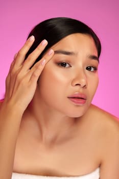 A young woman gently touches her face with one hand while showcasing a natural makeup look. The vibrant pink background adds a playful touch to her skincare routine.