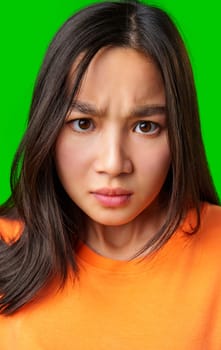 A young woman looks intensely at the camera with a serious expression. She has long black hair and is wearing an orange shirt, creating a vibrant contrast with the green backdrop.