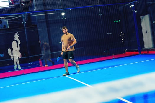 A young man engages in a friendly game of padel tennis on a striking blue court in an indoor sports facility, showcasing his skills and enthusiasm for the sport.