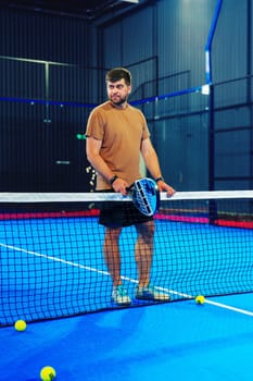 A male player stands at the net of a paddle tennis court, holding a paddle and looking ready for action. Bright blue court surface enhances the atmosphere, with balls nearby.