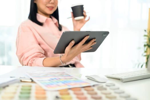 Female designer drinking coffee and using digital tablet at workplace.
