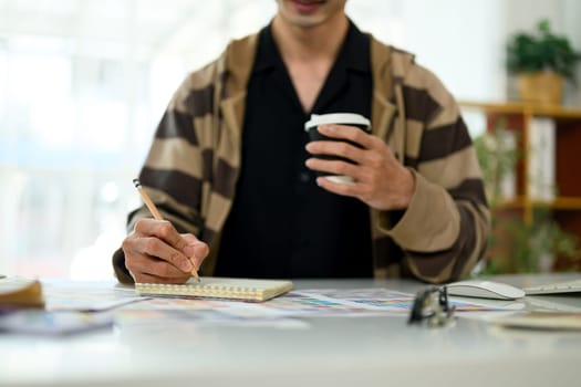 Creative man taking notes in a notebook and sipping coffee at workplace.