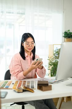 Young creative woman sitting at modern workspace checking project updates on smartphone.