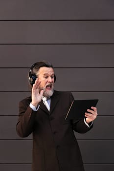 happy mature male in formal suit and wireless headphones using tablet for an online video call against gray wall, concept of middle age business man and technology of communication, copyspace for text