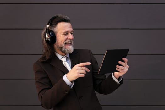 content mature male with beard in formal suit and wireless headphones using tablet while communicating online against gray wall, concept of middle age business man and technology of communication