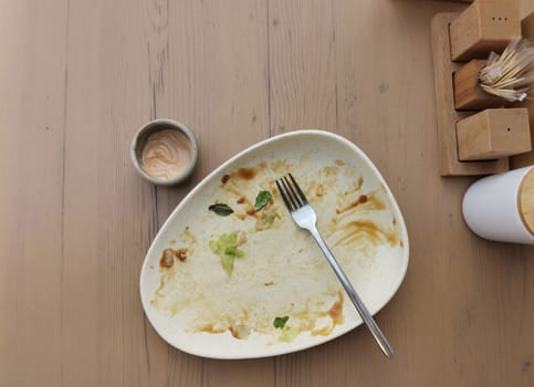Dirty plate after dinner. Top view of dirty white plate with fork over wooden tabletop. Dirty plate after eating salad with sauce on the dining table. Copy space