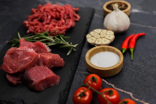 Side view of chunks of beef lying on a black cutting board, separated by sprigs of rosemary with red minced meat, surrounded by vegetables and seasonings on a dark background, close-up. The concept of cooking, shredding meat, diet, natural products. High quality photo