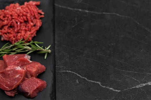 Top left view of chunks of beef lying on a black cutting board, separated by sprigs of rosemary with red minced meat on a dark background with space for text. The concept of cooking, shredding meat, diet, natural products. High quality photo