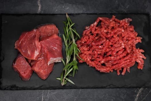 A close-up top view in the center of chunks of beef and minced meat, separated by sprigs of rosemary, are lying on a black cutting board on the left against a dark background with space for text. The concept of cooking, shredding meat, diet, natural products. High quality photo