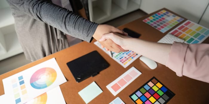 A close-up shot of graphic designers shaking hands, surrounded by color palettes and design materials on a collaborative workspace desk.