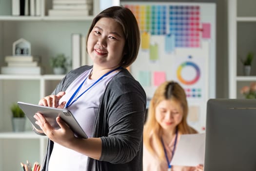 A graphic designer smiling at the camera while holding a tablet, working alongside a colleague in a colorful creative workspace.