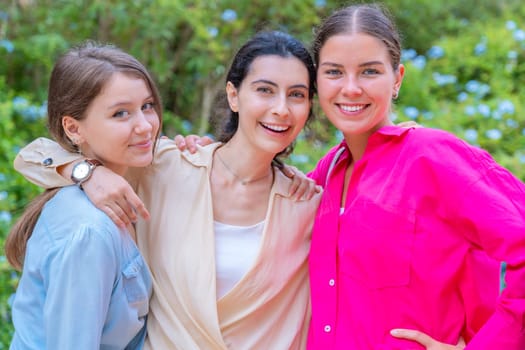 Three young women smiling and embracing each other while posing in a lush green park. They wear stylish casual outfits, expressing happiness, friendship, and joy in a natural outdoor setting.