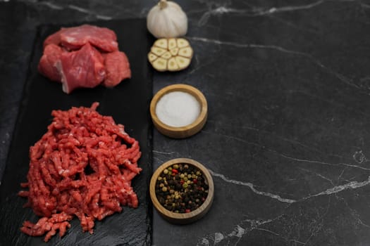 Top view of chunks of beef meat and red minced meat with streaks of fat lying on a black background with spices and salt in wooden bowls nearby and garlic. The concept of cooking, shredding meat, diet, natural products. High quality photo