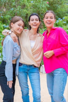 Three young women smiling and embracing each other while posing in a lush green park. They wear stylish casual outfits, expressing happiness, friendship, and joy in a natural outdoor setting.