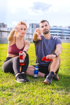 A fit man and woman sitting on the grass after a workout, holding water bottles and pointing at the camera. They appear relaxed and playful, enjoying a sunny outdoor setting in the city.