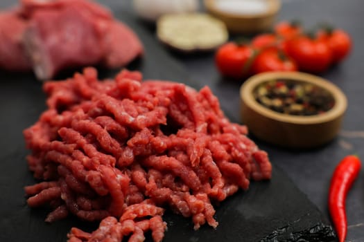 Close-up of raw red ground beef minced in a meat grinder lying on a black board, surrounded by out-of-focus cut meat, vegetables and seasonings in wooden bowls. The concept of diet, proper nutrition, natural products. High quality photo