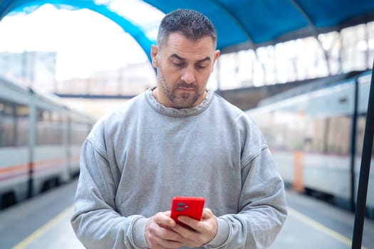 A cheerful man in a gray sweatshirt holds a red smartphone while standing at a train station. The urban setting conveys themes of travel, commuting, and digital connectivity.