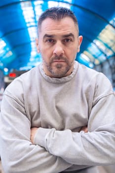 man in a gray sweatshirt standing at a train station with arms crossed. He has a determined expression, with a blue steel structure in the background. Urban travel and confidence concept.Vertical