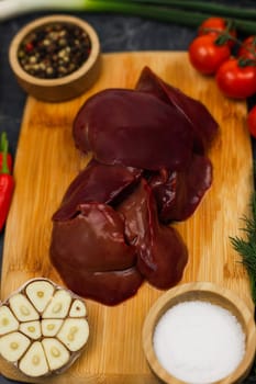 Top view, vertical close-up photo of poultry liver in the center lying on a wooden cutting board surrounded by vegetables, spices in wooden bowls and herbs. The concept of proper and healthy nutrition, vitamins and trace elements for the body, organics. High quality photo