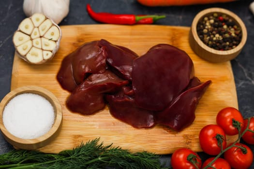 A close-up view of poultry liver from above in the center lies on a wooden cutting board, surrounded by vegetables, spices in wooden bowls, and herbs. The concept of proper and healthy nutrition, vitamins and trace elements for the body, organics. High quality photo