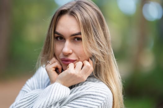 Beautiful blonde woman posing in a park in autumn, wearing a white woolen sweater, holding her neck with her hands