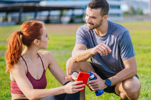 A young fit couple in sportswear relaxing after exercise in a green park. The woman is sitting on the grass using her smartphone, while the man crouches beside her with a water bottle, both smiling.