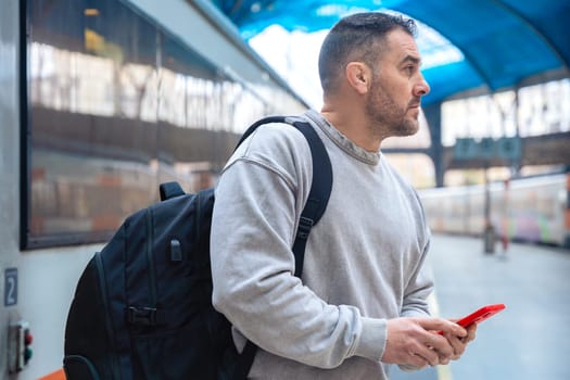 Serious man wearing a gray sweatshirt holding a red smartphone while looking around at a train station. His expression conveys anticipation, travel, and modern connectivity.