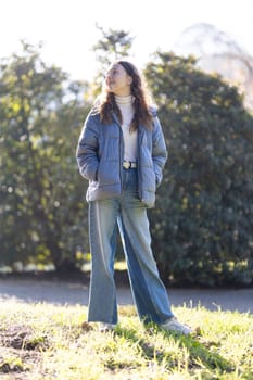Fashionable young woman standing in a park, enjoying the sunlight and looking up at the sky