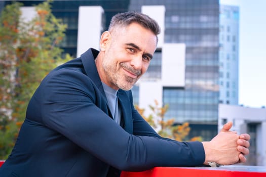 A middle-aged businessman in a stylish suit leaning on a railing and smiling. The modern city background enhances his confident, approachable, and professional demeanor.
