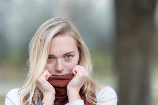 Young blonde woman pulling up her scarf over her mouth, feeling cold in a park during autumn