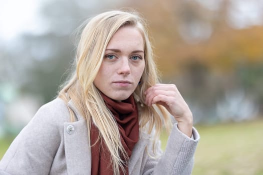 Young blonde woman wearing coat and scarf touching her hair while posing in a park during autumn