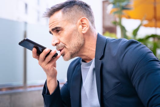 A confident businessman in a suit speaks into his smartphone using voice command. Seated in an outdoor urban setting, he looks focused and engaged in a professional conversation.