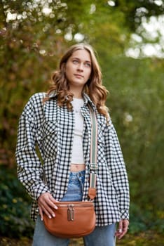 A young woman with wavy brown hair and blue eyes confidently poses outdoors in a lush autumn park. She wears a trendy black-and-white checkered shirt over a white crop top and classic blue jeans, accessorized with a tan leather crossbody bag with a striped strap. The soft natural lighting and blurred background of green foliage create a dreamy and cozy fall atmosphere. This fashionable portrait is ideal for lifestyle branding, autumn fashion campaigns, social media promotions, and advertising seasonal collections.
