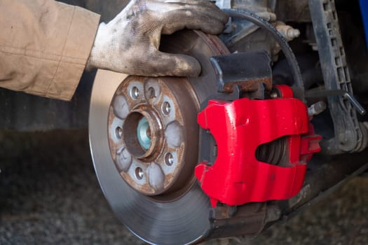 A person is working on a car's brake system, specifically the brake caliper. The caliper is red and has a black handle. The person is wearing gloves while working on the car