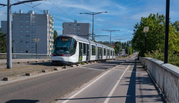 A tramway is traveling down a street of Strasbourg. The tramway is white and has a green stripe on it. The street is empty and the sky is blue