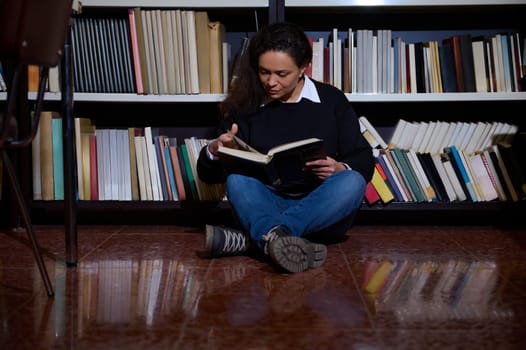 A young woman enjoys reading a book while sitting on the floor of a library. Bookshelves filled with various titles surround her, creating a cozy and contemplative atmosphere.
