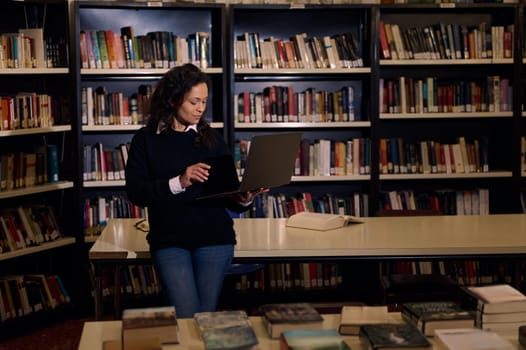 A woman stands in a library using a laptop for research. She is surrounded by bookshelves and a table with open and closed books, creating an atmosphere of knowledge and focus.