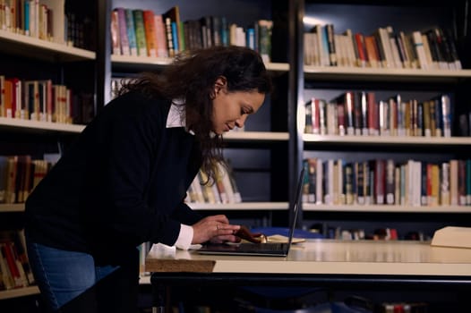 A dedicated woman stands studying on a laptop in a library, surrounded by shelves filled with books. The scene highlights focus, knowledge, and the pursuit of education.