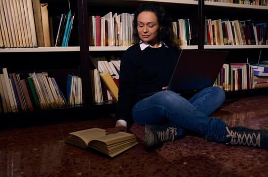 A woman seated on the floor of a library studies with a laptop and an open book, surrounded by shelves filled with books. The setting evokes themes of education, learning, and knowledge.