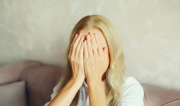 Upset sad crying woman covering her eyes and face with her hands while experiencing mental or physical suffering in room at home