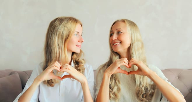Happy smiling middle aged mother with adult daughter together showing heart shaped gesture sign with hands and looking at each other at home