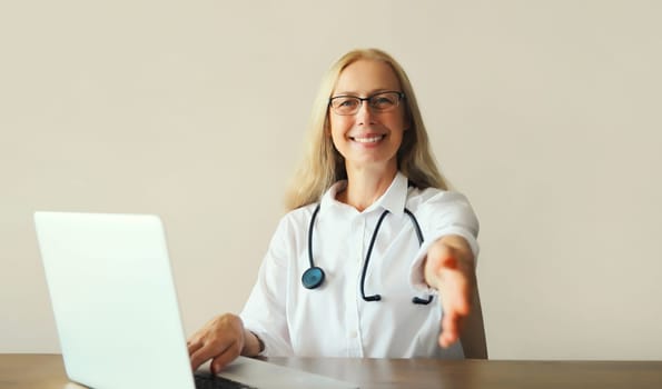 Happy middle-aged woman doctor working with laptop computer while sitting at desk in medical clinic office