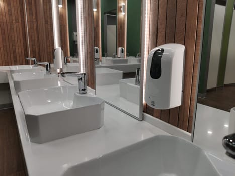 Liquid soap dispenser on wall in modern stylish public toilet interior. Automatic cleaning, washing hands using automatic sanitizer dispenser. Modern washroom interior with led-light on mirrors