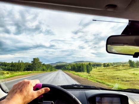 view through car windshield of nature landscape with green field, mountains, white clouds. Hand of woman on steering wheel. Female traveler